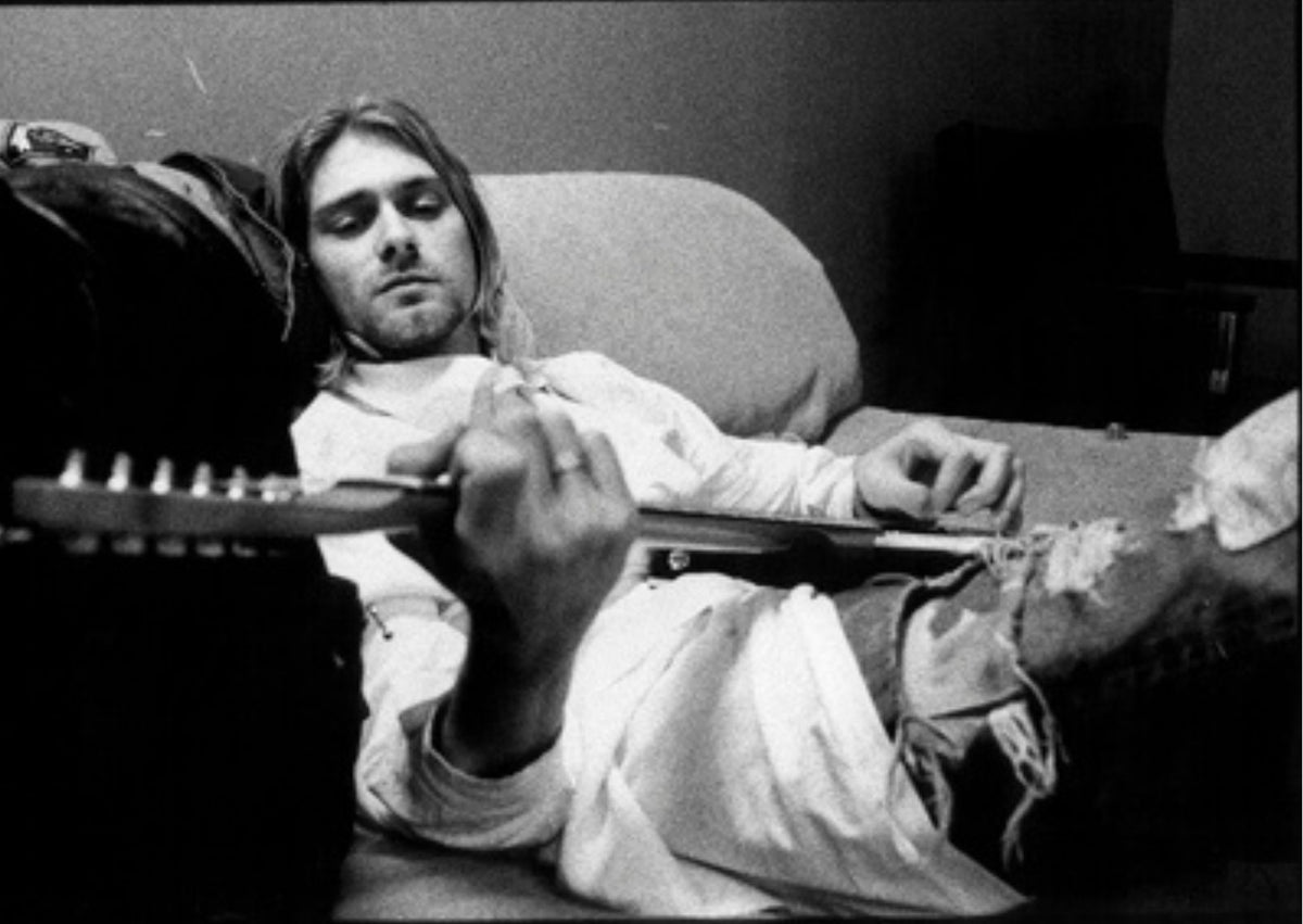 Pictured is Kurt Cobain of Nirvana in black and white film, sitting leisurely on a sofa playing his electric guitar.
