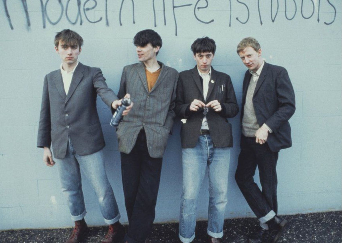 Pictured is all four members of Blur, with Damon holding a can of spray paint (far left) with all standing in front of a pale blue wall with text above their heads, which reads "modern life is rubbish".
