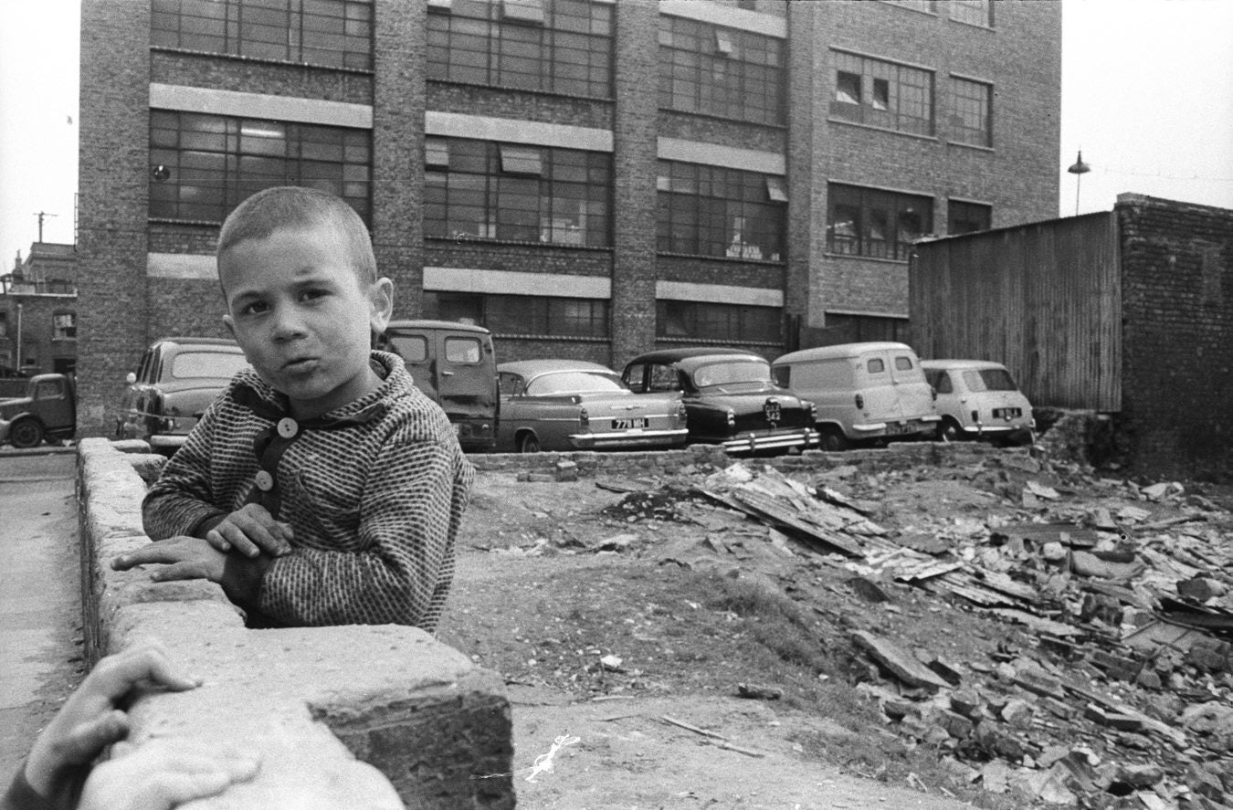 A little boy with a shaved head leaning over a wall in front of a bomb site
