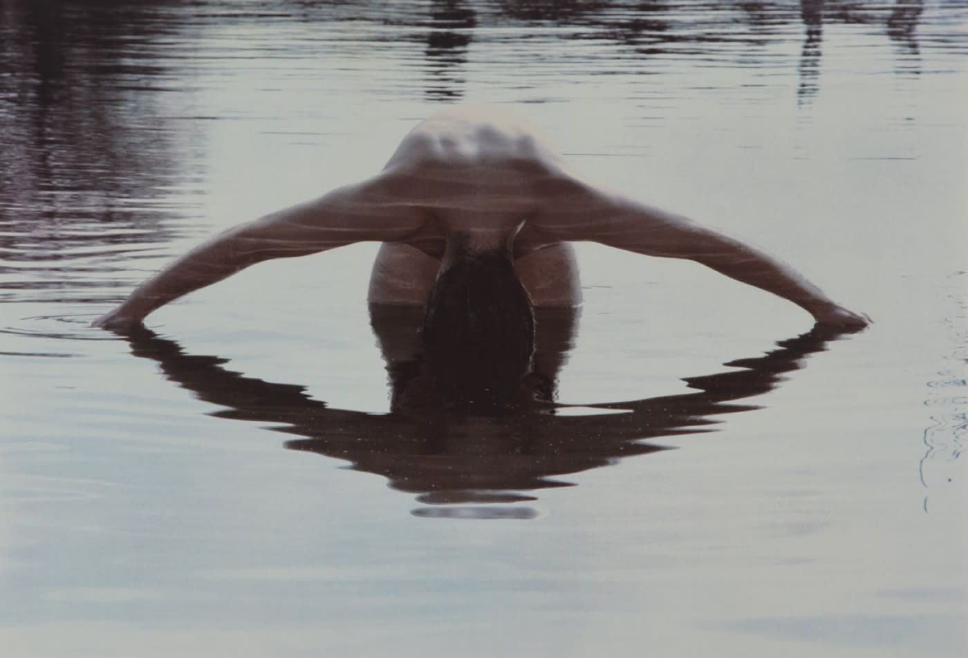 A woman dipping her hands in water whilst spread apart, with her body hunched together so the reflection mirrors her and creates an illusion of an eye