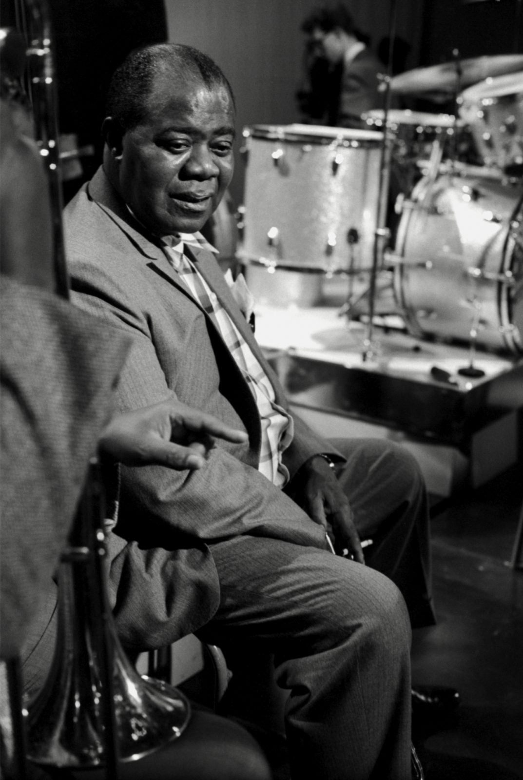 Legendary Louis Armstrong sitting grinning idly next besides a stage with a silver drum kit set up 