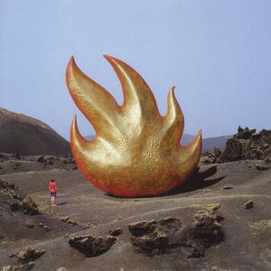 a man stood alone in a bare area of sandy textured ground with a giant statue of a flame towering over him 