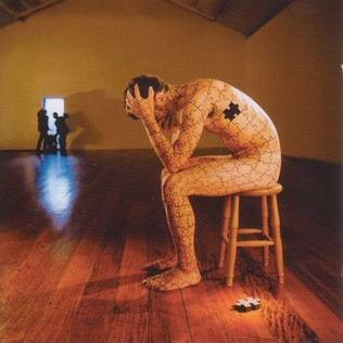 A naked man sitting on a chair, he is made out of puzzles and one piece is still on the floor. he has his head in his hands and there are people in the doorway just leaving