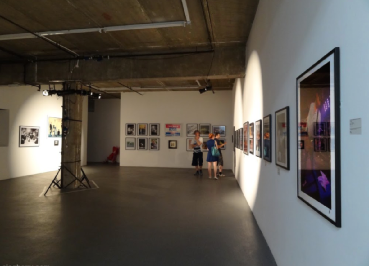 Pictured is the exhibition room with framed photos and graphic designs of the band, with white strobe lights, and three people admiring them.