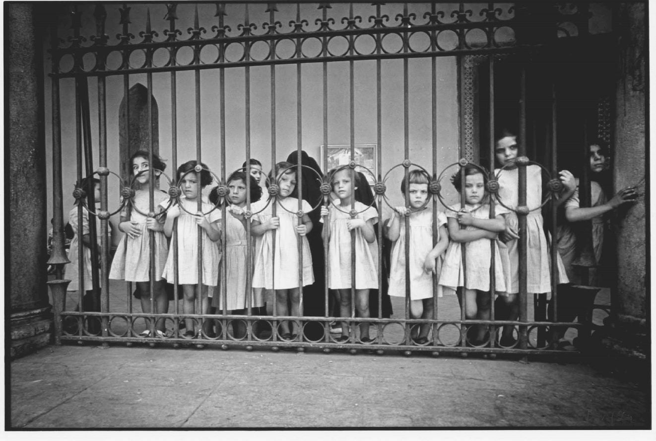 A group of little girls with their arms wrapped around the bars of a fence they are behind