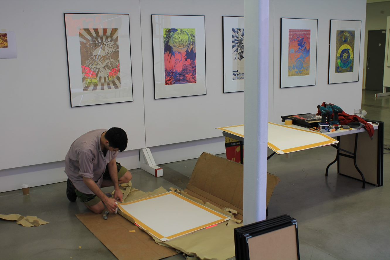 A man prepping one of the prints to go on the wall where five others are already up and in position 