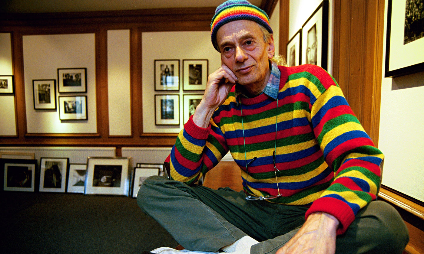 John "hoppy" Hopkins sitting on a desk with his legs crossed in a vibrant striped red yellow and blue jumper, holding his chin in one hand 